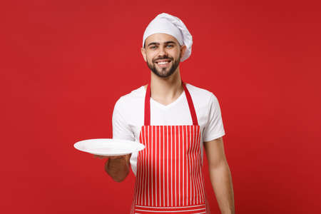Smiling young male chef cook or baker man in striped apron toque chefs hat isolated on red background. Cooking food concept. Mock up copy space. Holding empty blank round plate with place for food.の写真素材