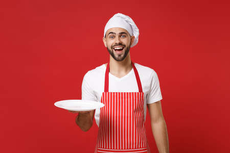 Cheerful young male chef cook or baker man in striped apron toque chefs hat isolated on red background. Cooking food concept. Mock up copy space. Holding empty blank round plate with place for food.の写真素材