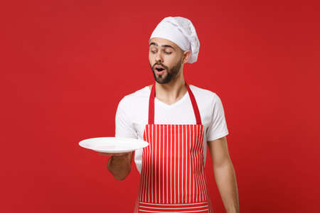 Shocked young male chef cook or baker man in striped apron toque chefs hat isolated on red background. Cooking food concept. Mock up copy space. Holding empty blank round plate with place for food.の写真素材