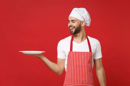 Cheerful young male chef cook or baker man in striped apron toque chefs hat isolated on red background. Cooking food concept. Mock up copy space. Holding empty blank round plate with place for food.の写真素材