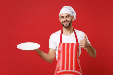 Smiling male chef cook or baker man in striped apron toque chefs hat isolated on red background. Cooking food concept. Mock up copy space. Hold empty blank plate with place for food showing thumb up.の写真素材