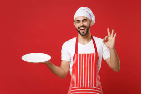 Joyful male chef cook or baker man in striped apron toque chefs hat isolated on red background. Cooking food concept. Mock up copy space. Hold empty blank plate with place for food showing OK gesture.の写真素材
