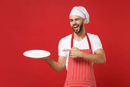 Cheerful chef cook or baker man in striped apron toque chefs hat isolated on red background. Cooking food concept. Mock up copy space. Pointing index finger on empty blank plate with place for food.の写真素材