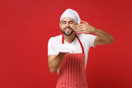 Chef cook or baker man in striped apron toque chefs hat isolated on red background. Cooking food concept. Mock up copy space. Holding plate with place for food raised hand to face, feeling food smell.の写真素材