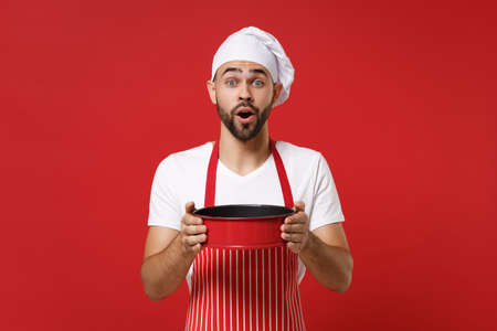 Shocked young bearded male chef cook or baker man in striped apron toque chefs hat posing isolated on red wall background. Cooking food concept. Mock up copy space. Holding metal baking form for pie.の写真素材