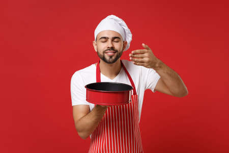 Chef cook or baker man in striped apron toque chefs hat posing isolated on red background. Cooking food concept. Mock up copy space. Hold baking form for pie, raised hand to face, feeling food smell.の写真素材