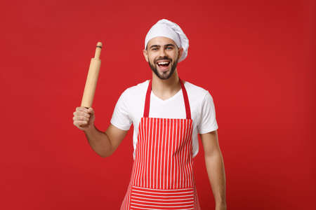 Laughing young bearded male chef cook or baker man in striped apron white t-shirt toque chefs hat posing isolated on red background. Cooking food concept. Mock up copy space. Hold wooden rolling pin.の写真素材