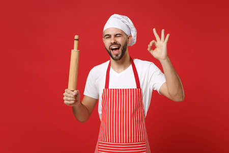 Bearded male chef cook or baker man in striped apron white t-shirt toque chefs hat posing isolated on red background. Cooking food concept. Mock up copy space. Holding rolling pin, showing OK gesture.の写真素材