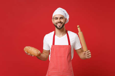 Smiling young bearded male chef cook or baker man in striped apron white t-shirt toque chefs hat posing isolated on red background. Cooking food concept. Mock up copy space. Hold rolling pin, bread.の写真素材