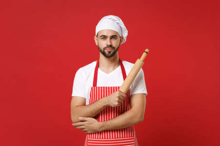 Handsome young bearded male chef cook or baker man in striped apron white t-shirt toque chefs hat posing isolated on red background. Cooking food concept. Mock up copy space. Hold wooden rolling pin.の写真素材