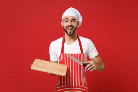 Excited young bearded male chef cook or baker man in striped apron white t-shirt toque chefs hat posing isolated on red background. Cooking food concept. Mock up copy space. Hold knife, cutting board.の写真素材