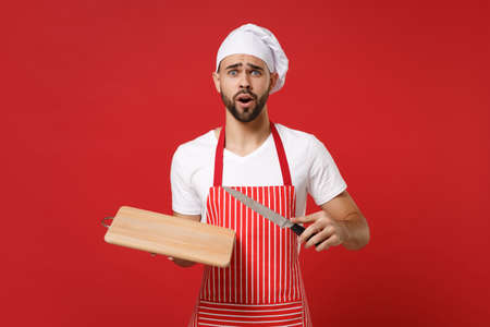Shocked young bearded male chef cook or baker man in striped apron white t-shirt toque chefs hat posing isolated on red background. Cooking food concept. Mock up copy space. Hold knife, cutting board.の写真素材