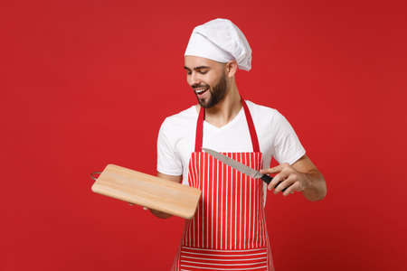 Handsome young bearded male chef cook or baker man in striped apron white t-shirt toque chefs hat posing isolated on red background. Cooking food concept. Mock up copy space. Hold knife cutting board.の写真素材