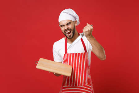 Crazy young bearded male chef cook or baker man in striped apron white t-shirt toque chefs hat posing isolated on red background. Cooking food concept. Mock up copy space. Holding knife cutting board.の写真素材