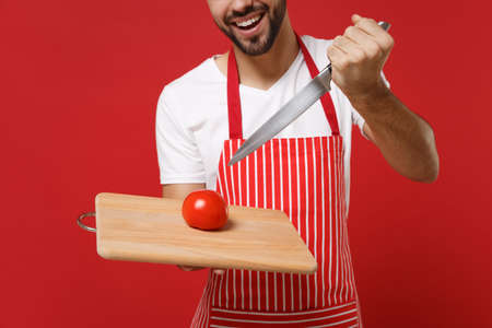Cropped image of bearded male chef cook or baker man in striped apron white t-shirt posing isolated on red background. Cooking food concept. Mock up copy space. Hold knife cutting board, fresh tomato.の写真素材