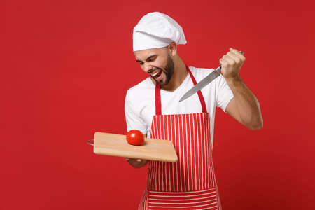 Crazy young male chef cook or baker man in striped apron toque chefs hat posing isolated on red background. Cooking food concept. Mock up copy space. Hold knife cutting board, fresh tomato, screaming.の写真素材