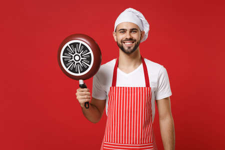 Smiling young bearded male chef cook or baker man in striped apron white t-shirt toque chefs hat posing isolated on red wall background. Cooking food concept. Mock up copy space. Holding frying pan.の写真素材