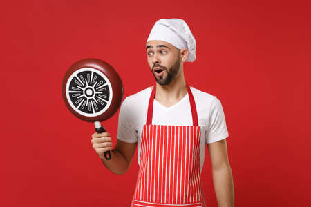 Shocked young bearded male chef cook or baker man in striped apron white t-shirt toque chefs hat posing isolated on red wall background. Cooking food concept. Mock up copy space. Holding frying pan.の写真素材