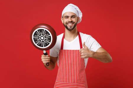 Smiling young male chef cook or baker man in striped apron white t-shirt toque chefs hat posing isolated on red background. Cooking food concept. Mock up copy space. Hold frying pan, showing thumb up.の写真素材