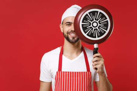 Smiling young male chef cook or baker man in striped apron white t-shirt toque chefs hat posing isolated on red background. Cooking food concept. Mock up copy space. Covering face with frying pan.の写真素材