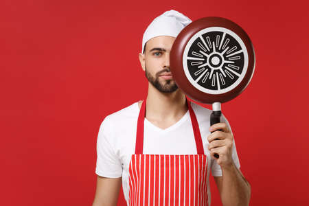 Bearded young male chef cook or baker man in striped apron white t-shirt toque chefs hat posing isolated on red background. Cooking food concept. Mock up copy space. Covering face with frying pan.の写真素材