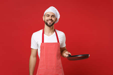 Handsome young bearded male chef cook or baker man in striped apron white t-shirt toque chefs hat posing isolated on red wall background. Cooking food concept. Mock up copy space. Holding frying pan.の写真素材