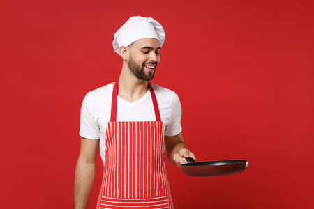 Smiling young bearded male chef cook or baker man in striped apron white t-shirt toque chefs hat posing isolated on red wall background. Cooking food concept. Mock up copy space. Holding frying pan.の写真素材