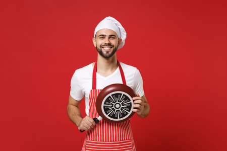 Smiling young bearded male chef cook or baker man in striped apron white t-shirt toque chefs hat posing isolated on red wall background. Cooking food concept. Mock up copy space. Holding frying pan.の写真素材