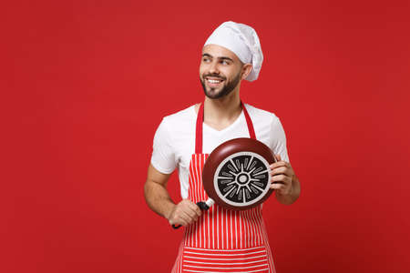Smiling young bearded male chef cook or baker man in striped apron toque chefs hat posing isolated on red wall background. Cooking food concept. Mock up copy space. Holding frying pan, looking aside.の写真素材