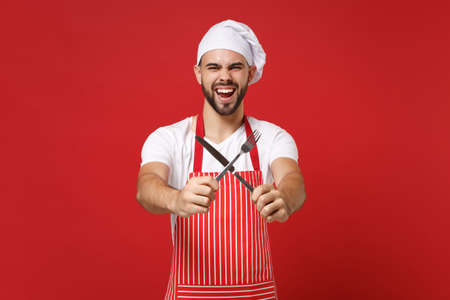 Screaming young bearded male chef cook or baker man in striped apron toque chefs hat posing isolated on red background. Cooking food concept. Mock up copy space. Hold cutlery fork and knife crossed.の写真素材