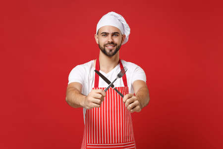 Smiling young bearded male chef cook or baker man in striped apron toque chefs hat posing isolated on red background. Cooking food concept. Mock up copy space. Holding cutlery fork and knife crossed.の写真素材