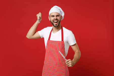 Crazy young bearded male chef cook or baker man in striped apron toque chefs hat posing isolated on red background. Cooking food concept. Mock up copy space. Holding cutlery fork and knife, screaming.の写真素材