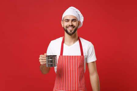 Smiling young bearded male chef cook or baker man in striped apron toque chefs hat posing isolated on red background. Cooking food concept. Mock up copy space. Holding metal iron sieve sifting flour.の写真素材
