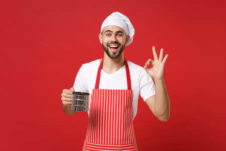 Young male chef cook or baker man in striped apron toque chefs hat posing isolated on red background. Cooking food concept. Mock up copy space. Hold metal iron sieve sifting flour, showing OK gesture.の写真素材