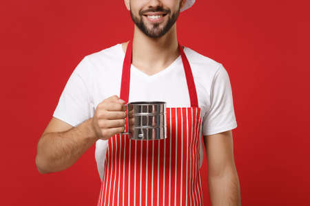 Cropped image of bearded male chef cook or baker man in striped apron white t-shirt posing isolated on red background. Cooking food concept. Mock up copy space. Holding metal iron sieve sifting flour.の写真素材