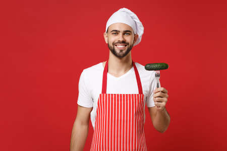 Smiling young bearded male chef cook or baker man in striped apron toque chefs hat posing isolated on red wall background. Cooking food concept. Mock up copy space. Holding fork with fresh cucumber.の写真素材