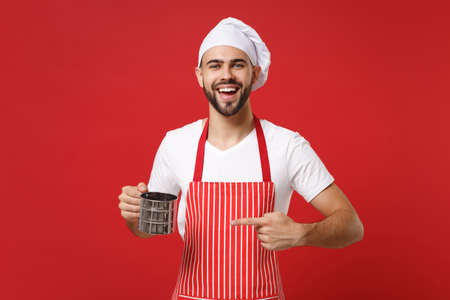 Young male chef cook or baker man in striped apron toque chefs hat posing isolated on red background. Cooking food concept. Mock up copy space. Pointing index finger on metal iron sieve sifting flour.の写真素材