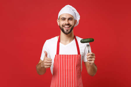 Smiling bearded male chef cook or baker man in striped apron toque chefs hat posing isolated on red background. Cooking food concept. Mock up copy space. Holding fork with cucumber, showing thumb up.の写真素材