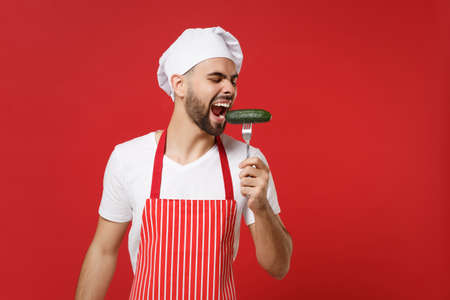 Crazy young bearded male chef cook or baker man in striped apron toque chefs hat posing isolated on red background in studio. Cooking food concept. Mock up copy space. Biting fresh cucumber on fork.の写真素材