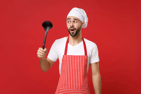 Confused young bearded male chef cook or baker man in striped apron toque chefs hat posing isolated on red wall background. Cooking food concept. Mock up copy space. Holding soup black ladle dipper.の写真素材