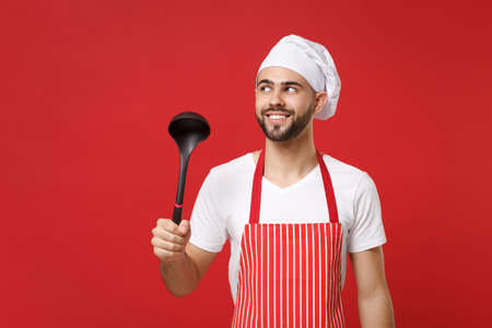 Smiling young bearded male chef cook or baker man in striped apron toque chefs hat posing isolated on red wall background. Cooking food concept. Mock up copy space. Holding soup black ladle dipper.の写真素材