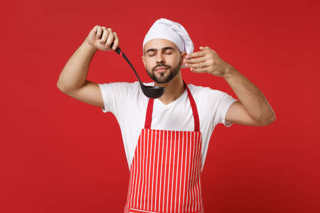 Chef cook baker man in striped apron toque chefs hat posing isolated on red background. Cooking food concept. Mock up copy space. Hold soup black ladle dipper raised hand to face feeling food smell.の写真素材