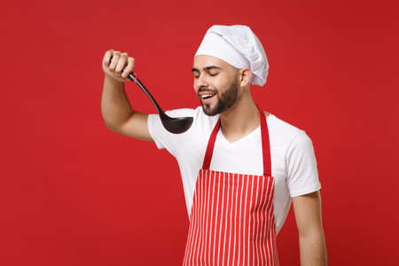 Young bearded male chef cook or baker man in striped apron toque chefs hat posing isolated on red wall background. Cooking food concept. Mock up copy space. Hold soup black ladle dipper, tasting food.の写真素材