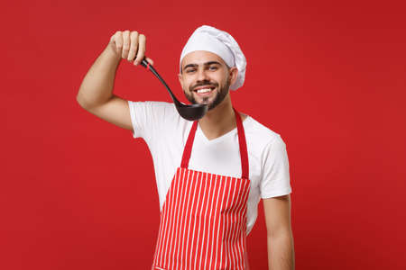 Smiling young bearded male chef cook or baker man in striped apron toque chefs hat posing isolated on red wall background. Cooking food concept. Mock up copy space. Holding soup black ladle dipper.の写真素材