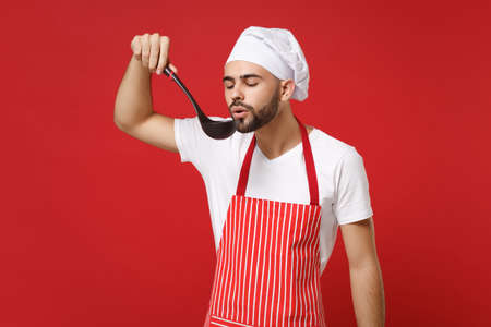 Young bearded male chef cook or baker man in striped apron toque chefs hat posing isolated on red wall background. Cooking food concept. Mock up copy space. Hold soup black ladle dipper, tasting food.の写真素材