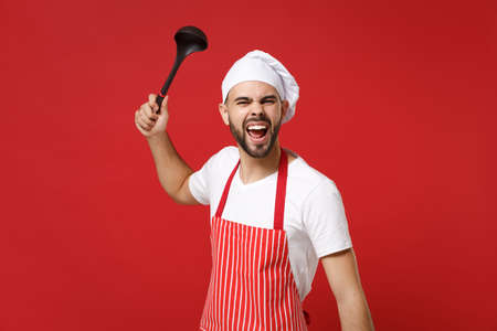 Crazy young bearded male chef cook or baker man in striped apron toque chefs hat posing isolated on red background. Cooking food concept. Mock up copy space. Holding soup black ladle dipper screaming.の写真素材
