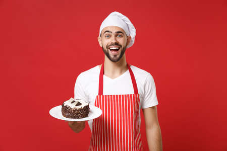 Excited young bearded male chef cook or baker man in striped apron white t-shirt toque chefs hat posing isolated on red background. Cooking food concept. Mock up copy space. Holding plate with cake.の写真素材