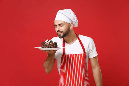 Young male chef cook or baker man in striped apron white t-shirt toque chefs hat posing isolated on red background. Cooking food concept. Mock up copy space. Hold plate with cake, feeling food smell.の写真素材