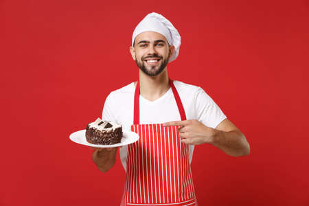 Smiling male chef cook or baker man in striped apron white t-shirt toque chefs hat posing isolated on red background. Cooking food concept. Mock up copy space. Point index finger on plate with cake.の写真素材