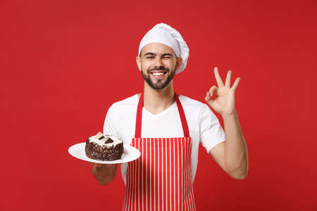 Bearded male chef cook or baker man in striped apron white t-shirt toque chefs hat posing isolated on red background. Cooking food concept. Mock up copy space. Hold plate with cake showing OK gesture.の写真素材